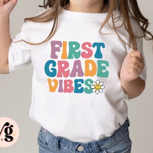 Peut inclure: T-shirt blanc avec le texte "First Grade Vibes" en police r&eacute;tro color&eacute;e. Une marguerite jaune avec un visage souriant est &agrave; c&ocirc;t&eacute; du texte.