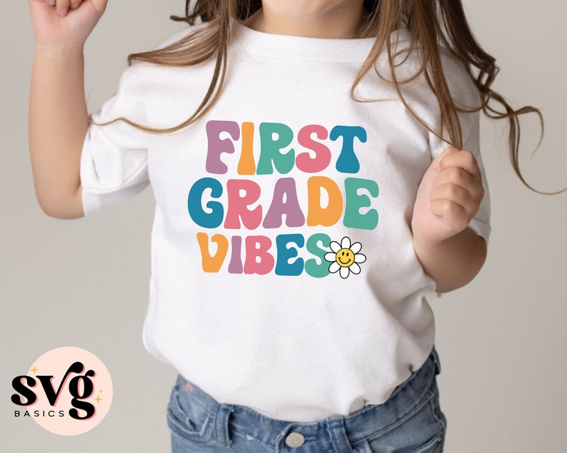 Peut inclure: T-shirt blanc avec le texte "First Grade Vibes" en police r&eacute;tro color&eacute;e. Une marguerite jaune avec un visage souriant est &agrave; c&ocirc;t&eacute; du texte.