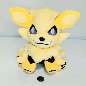 May include: A plush toy in the shape of a yellow and cream-colored creature with large ears and blue eyes. The toy has black accents on its paws and a small coin is next to it.