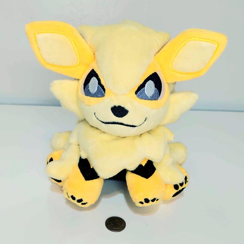 May include: A plush toy in the shape of a yellow and cream-colored creature with large ears and blue eyes. The toy has black accents on its paws and a small coin is next to it.