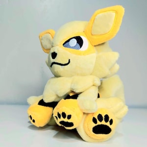 May include: A plush toy of a yellow and cream-colored creature with large ears, blue eyes, and black paw prints. The toy has a friendly expression and is made of soft, fluffy material. The toy is a collectible item.