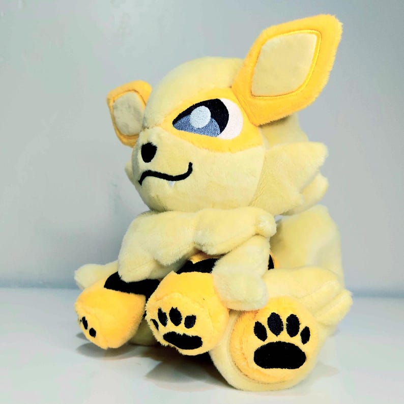 May include: A plush toy of a yellow and cream-colored creature with large ears, blue eyes, and black paw prints. The toy has a friendly expression and is made of soft, fluffy material. The toy is a collectible item.