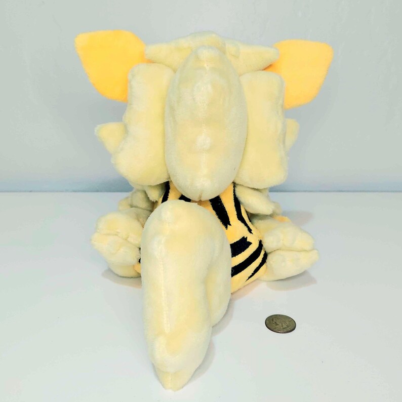 May include: A plush toy in a seated position, featuring a light yellow body with orange ears. The toy has a black and yellow striped pattern on its belly. A coin is visible near the toy.