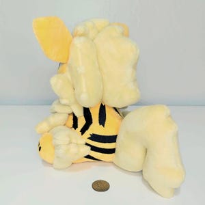 May include: A plush toy tiger in yellow and black stripes. The toy has large, floppy ears and a rounded body. The tiger is sitting down, with its legs extended. A penny is in front of the toy.