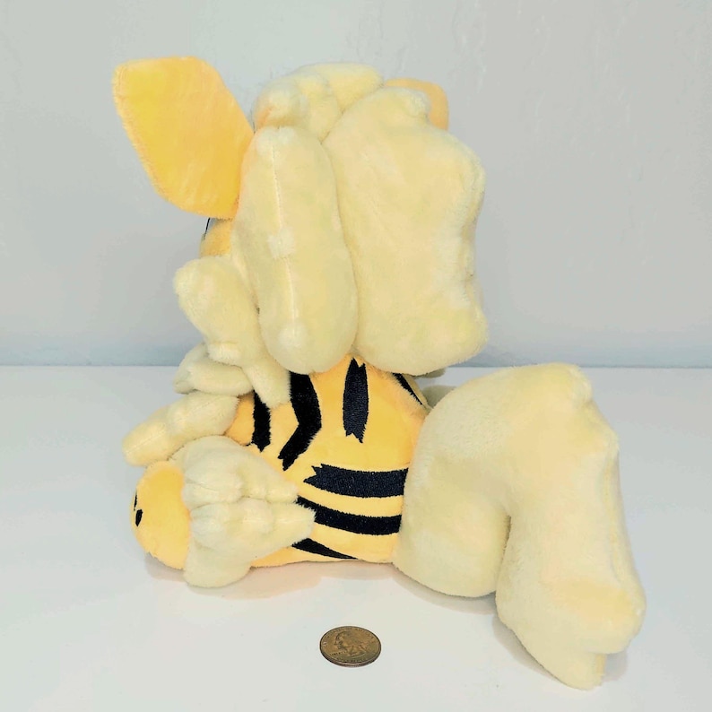 May include: A plush toy tiger in yellow and black stripes. The toy has large, floppy ears and a rounded body. The tiger is sitting down, with its legs extended. A penny is in front of the toy.