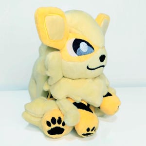 May include: A plush toy of a yellow and cream-colored creature with large ears, blue eyes, and black paw prints. The toy has a fluffy mane and black stripes on its legs. The overall design is soft and cuddly.