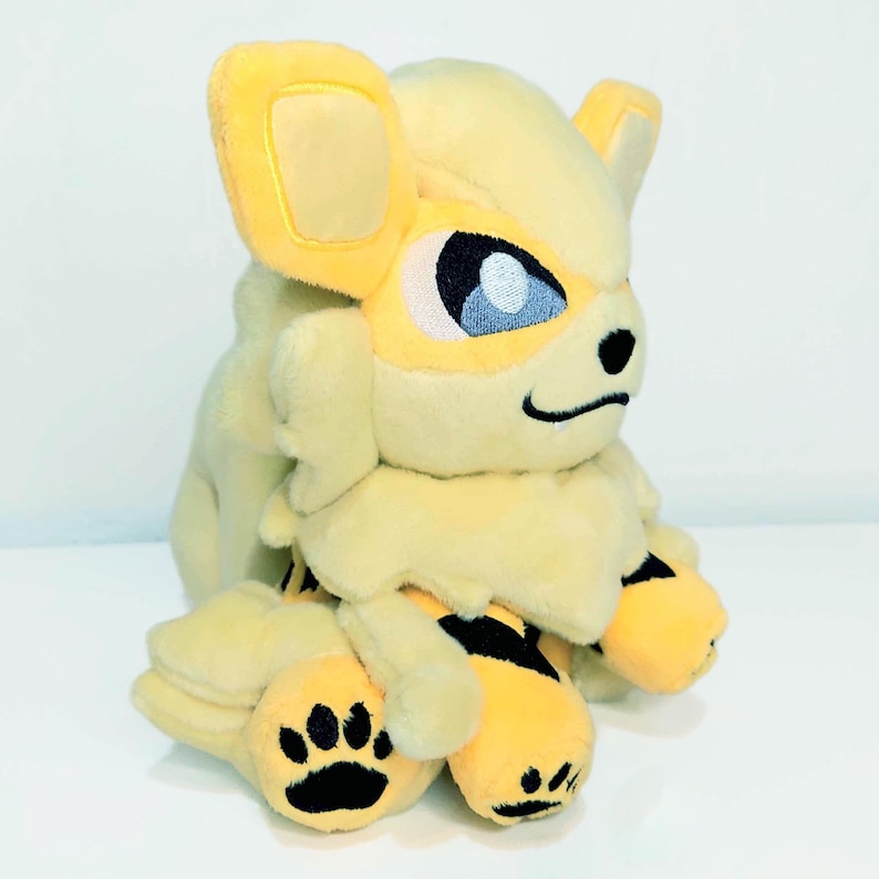 May include: A plush toy of a yellow and cream-colored creature with large ears, blue eyes, and black paw prints. The toy has a fluffy mane and black stripes on its legs. The overall design is soft and cuddly.