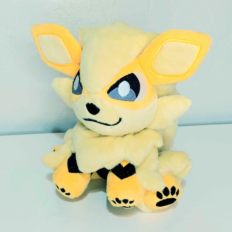 May include: A plush toy of a yellow and cream-colored creature with large ears, blue eyes, and black paw prints. The toy has a smiling face and black accents. The creature's paws and chest have black markings.