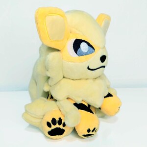May include: A plush toy of a yellow and cream-colored creature with large ears, blue eyes, and black paw prints. The toy has a fluffy mane and black stripes on its legs. The overall design is soft and cuddly.