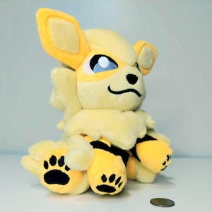 May include: A plush toy of a yellow and cream-colored creature with large ears, blue eyes, and black paw prints. The toy has a fluffy mane and tail, and is sitting down. A coin is in the foreground.