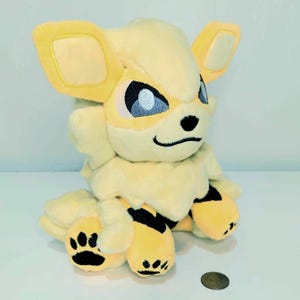 May include: A plush toy of a yellow and cream-colored creature with large ears, blue eyes, and black markings. The toy has a smiling expression and black paw prints on its feet. A coin is next to the toy.