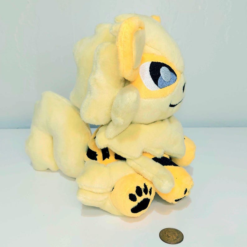 May include: A plush toy in the shape of a yellow and cream-colored creature with black accents. The toy has large blue eyes, a black paw print, and a smiling expression. The toy is sitting on a white surface.