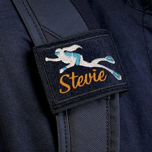 Op de afbeelding: Een rechthoekige zwarte stoffen patch bevestigd aan een donker marineblauwe riem. De patch bevat een geborduurde witte en lichtblauwe afbeelding van een duiker in beweging, geplaatst boven de geborduurde oranje cursieve tekst die Stevie leest.