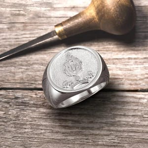 May include: A silver signet ring with a detailed engraved crest featuring a crown, shield, and floral accents.