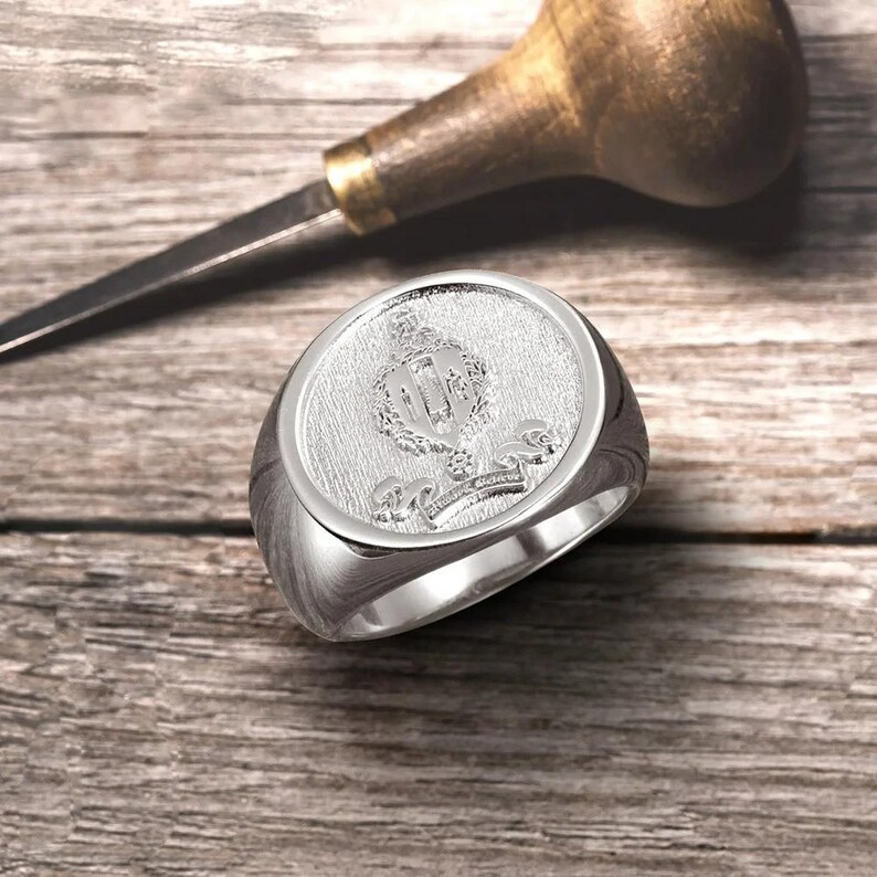 May include: A silver signet ring with a detailed engraved crest featuring a crown, shield, and floral accents.