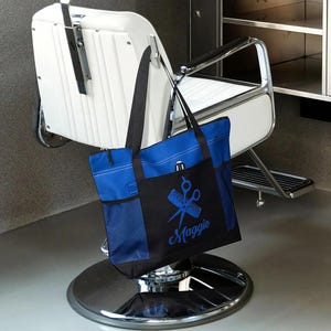 May include: A blue and black tote bag with a design of scissors and a comb, with the name "Maggie" embroidered in blue. The bag is sitting on a barber's chair.
