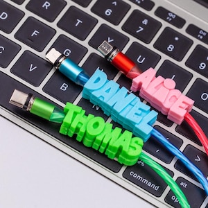 Puede incluir: Tres cables de carga con etiquetas de nombre impresas en 3D. Un cable verde tiene el nombre THOMAS, un cable azul tiene el nombre DANIEL y un cable rojo tiene el nombre ALICE. Cada cable tiene una punta de conector metálica.