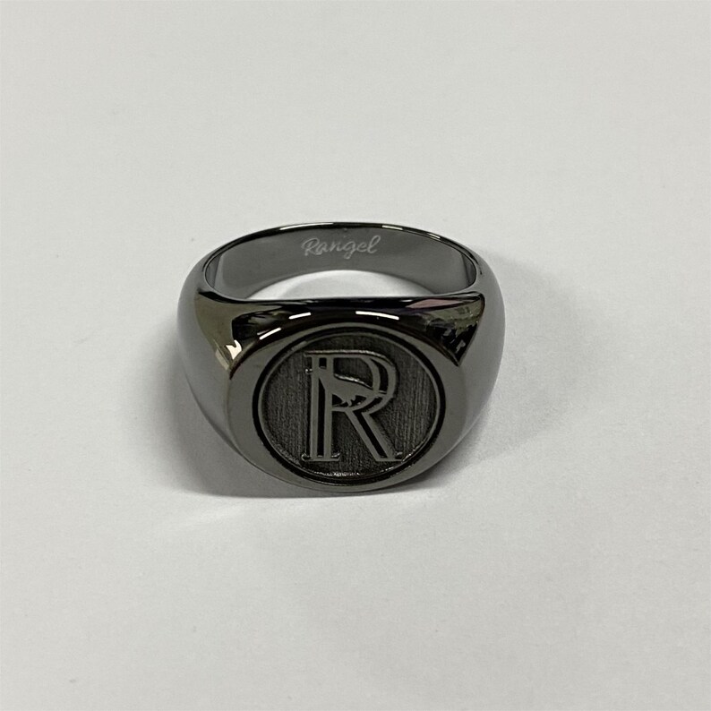 May include: A black signet ring with a raised, engraved letter "R" in the center. The ring is inscribed with the word "Rangel" on the inside band.