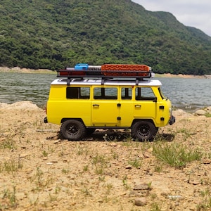 May include: A yellow toy off-road van with a white roof and black wheels. The van has a roof rack with accessories. The background features a lake and a green mountain.