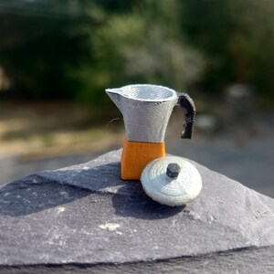 May include: A miniature stovetop espresso maker featuring a silver-colored upper chamber and lid, a bright orange base, and a black handle. The lid is detached and rests beside the main body of the coffee pot, showing a textured, layered finish.