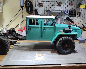 Può includere: Un camion giocattolo verde turchese con tetto bianco e dettagli neri. Il camion ha una grande cabina con quattro porte e un pianale di carico sul retro. Il camion ha ruote nere con dettagli rossi e si trova su una superficie di legno.