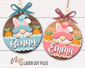 May include: Two wooden round ornaments with bunny-eared gnomes wearing hats. The ornament on the left is blue and says "Benny". The ornament on the right is pink and says "Emma".