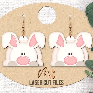 Puede incluir: Un par de pendientes de madera cortados con láser que presentan un diseño de conejo blanco con orejas y nariz rosas. Los pendientes cuelgan de un fondo marrón con el texto "Mrs. Laser Cut Files".