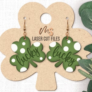 May include: A pair of green shamrock-shaped earrings with white polka dots and the word "lucky" written on each earring. The earrings are hanging from a brown display card.