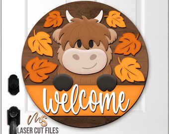 May include: A round wooden welcome sign featuring a cartoon Highland cow with brown fur, white horns, and a smiling face. Orange leaves surround the cow. The word "welcome" is written in white script on an orange banner.