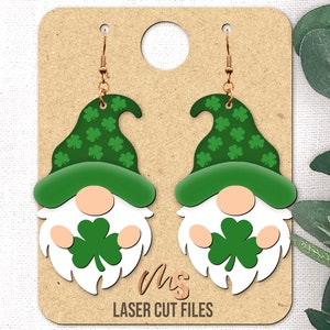 May include: A pair of green and white gnome earrings with shamrock designs. The earrings are made of wood and have a gold hook.