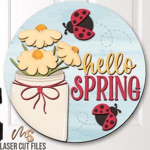 Può includere: Un cartello rotondo in legno con sfondo blu e venature del legno dipinte di bianco. Il cartello presenta un barattolo di vetro pieno di margherite gialle e coccinelle rosse. Il testo "hello spring" è scritto in lettere gialle e rosse.