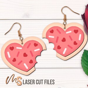 Könnte beinhalten: Ein Paar herzförmige Ohrringe mit einem rosa und weißen Zuckerguss-Design, das an einen angebissenen Keks erinnert. Die Ohrringe sind aus Holz und haben goldfarbene Haken.
