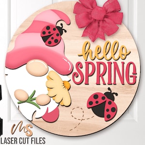 Puede incluir: Un letrero redondo de madera con un gnomo rosa y blanco que sostiene una flor amarilla. El letrero dice "hello spring" en letras amarillas y rosas. Hay dos mariquitas en el letrero, una con manchas rojas y negras y la otra con manchas rosas y negras.