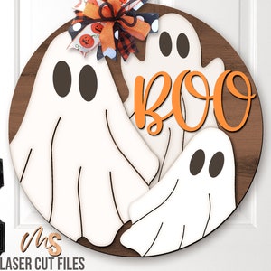 May include: A round wooden door hanger with three white ghosts and the word "BOO" in orange letters. The ghosts have black eyes and are layered on top of each other. A black and white checkered bow with orange pumpkins is at the top of the hanger.