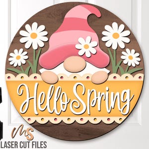 Può includere: Un cartello rotondo di legno con un gnomo rosa che indossa un cappello rosa e tiene uno striscione che dice "Hello Spring". Lo gnomo è circondato da margherite bianche con centri gialli.