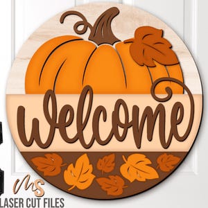 May include: A round wooden welcome sign featuring a large orange pumpkin with a brown stem and leaf. The word "Welcome" is in brown script, and the bottom border has autumn leaves. The sign is designed for fall and seasonal decor.