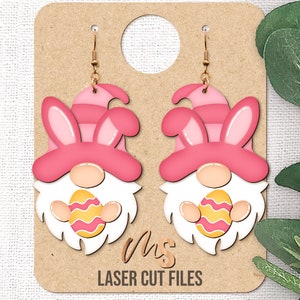 May include: A pair of pink and white Easter gnome earrings with bunny ears and holding an Easter egg. The earrings are on a brown card with the text "Ohs Laser Cut Files".