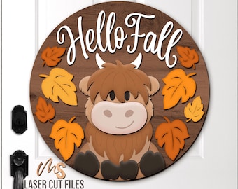 May include: A round wooden sign with a brown background and the words "Hello Fall" in white script. A cartoon Highland cow is centered, surrounded by orange and yellow fall leaves. The sign is a fall-themed decoration.
