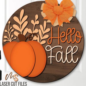 Pode incluir: Uma placa de porta redonda de madeira com tema de outono com uma abóbora e as palavras "Hello Fall". A placa apresenta uma abóbora laranja, folhagem de outono e um laço laranja. O fundo é um grão de madeira escura.