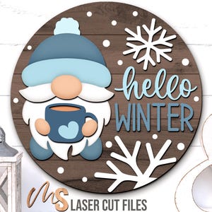 May include: A circular wooden sign featuring a winter gnome design. The gnome is wearing a blue hat and holding a blue mug with a heart. The words "hello WINTER" are in blue. White snowflakes and the text "MS LASER CUT FILES" are also visible.