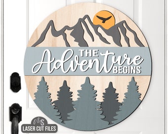 May include: A round wooden sign with the text "The Adventure Begins" in white lettering on a light blue background. The sign features a silhouette of mountains, a sun, and a bird flying overhead. There are also five dark grey trees at the bottom of the sign.