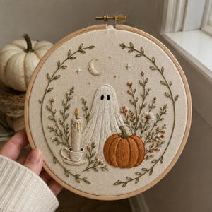 May include: A circular wooden embroidery hoop containing a fabric panel with hand-stitched designs. The embroidery features a white ghost, an orange pumpkin, a lit candle in a cup, and a crescent moon, all framed by delicate green leafy vines.