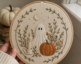 May include: A circular wooden embroidery hoop containing a fabric panel with hand-stitched designs. The embroidery features a white ghost, an orange pumpkin, a lit candle in a cup, and a crescent moon, all framed by delicate green leafy vines.