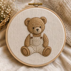 May include: A circular wooden embroidery hoop containing a piece of off-white fabric. Centered on the fabric is a detailed embroidery of a brown teddy bear wearing light beige overalls with a small front pocket and two shoulder straps.