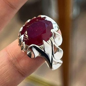 May include: A silver ring with a large, oval-shaped ruby gemstone. The ring has a detailed, ornate design.