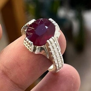 Puede incluir: Un anillo de plata con una gran piedra preciosa roja oscura engastada en una garra. El anillo tiene una banda texturizada.