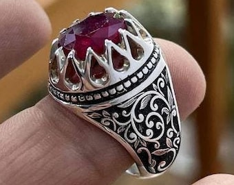 May include: A silver ring with a large, faceted, red gemstone. The ring has a detailed, black floral pattern on the band and a decorative crown-like setting around the stone. The ring is held up by a finger.