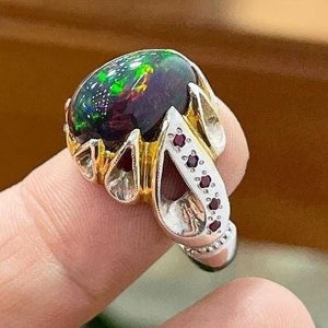 Op de afbeelding: Een zilveren ring met een grote, ovale zwarte opaal-edelsteen die in een gouden zetting is gezet. De ring is versierd met kleine rode edelstenen rond de band.