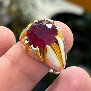 Op de afbeelding: Een goud-zilveren ring met een grote, gefacetteerde, dieprode edelsteen. De ring heeft een klassiek ontwerp met een gouden zetting rond de edelsteen en een zilveren band. De edelsteen is het middelpunt en vangt het licht.