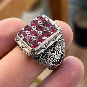 Könnte beinhalten: Ein silberner Ring mit rechteckiger Oberseite, besetzt mit zahlreichen kleinen, oval geformten roten Edelsteinen. Das Band des Rings weist komplizierte, dunkle Blumenmuster auf. Der Ring wird von einem Finger gehalten.